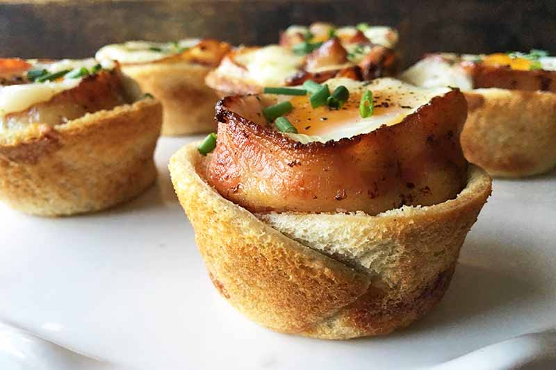 Horizontal image of baked toast cups with bacon, eggs, and chive garnish.