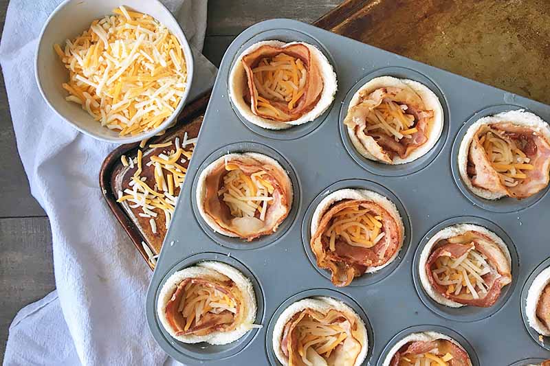 Horizontal image of a muffin tin lined with bread and bacon, with cheese in the center of each tin, and a bowl of shredded cheese on the side.