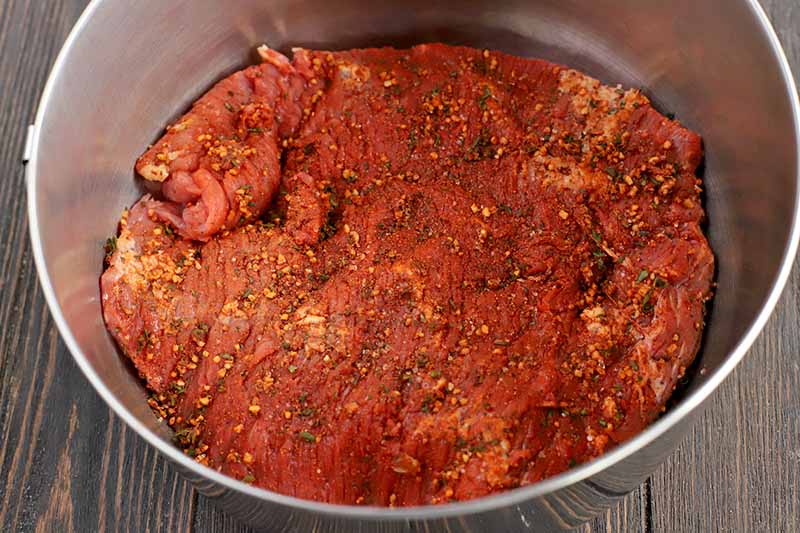 Raw beef coated in a red mixture of herbs and spices, in a large stainless steel bowl, on a dark brown wood table.