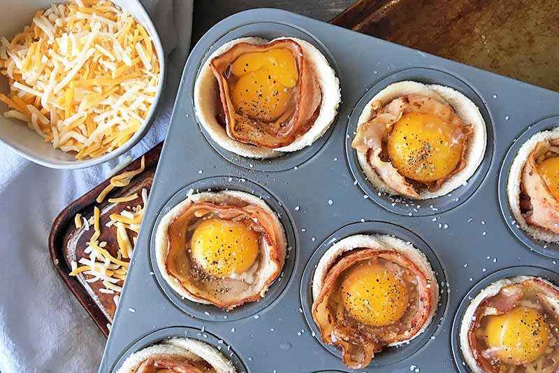 Horizontal image of a muffin tin with unbaked breakfast cups and a bowl of cheese on the side.
