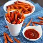 Horizontal image of a bowl and cup with orange fries and a cup of ketchup on a blue napkin.
