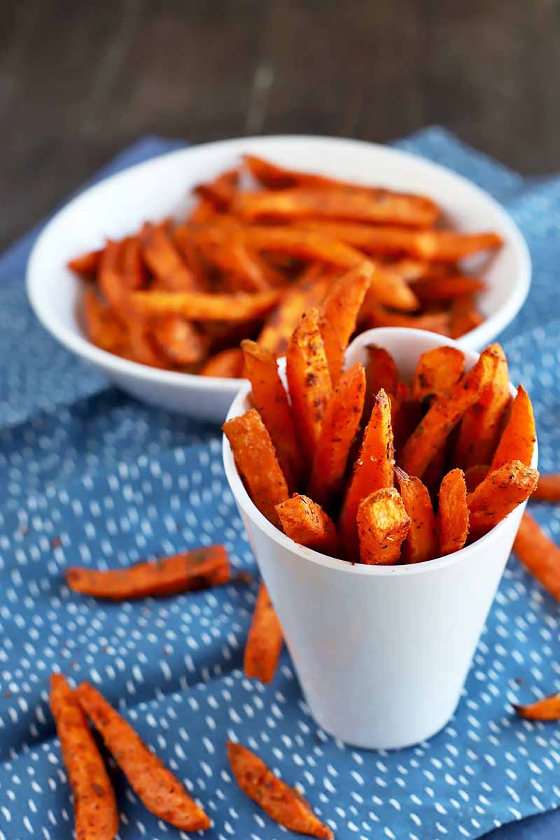 Vertical image of sweet potato fries in two separate dishes on a blue napkin.