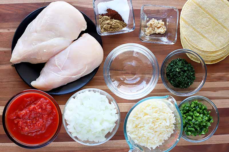 Horizontal image of assorted fresh herbs, cheese, cut vegetables, and chicken breasts on a wooden surface.