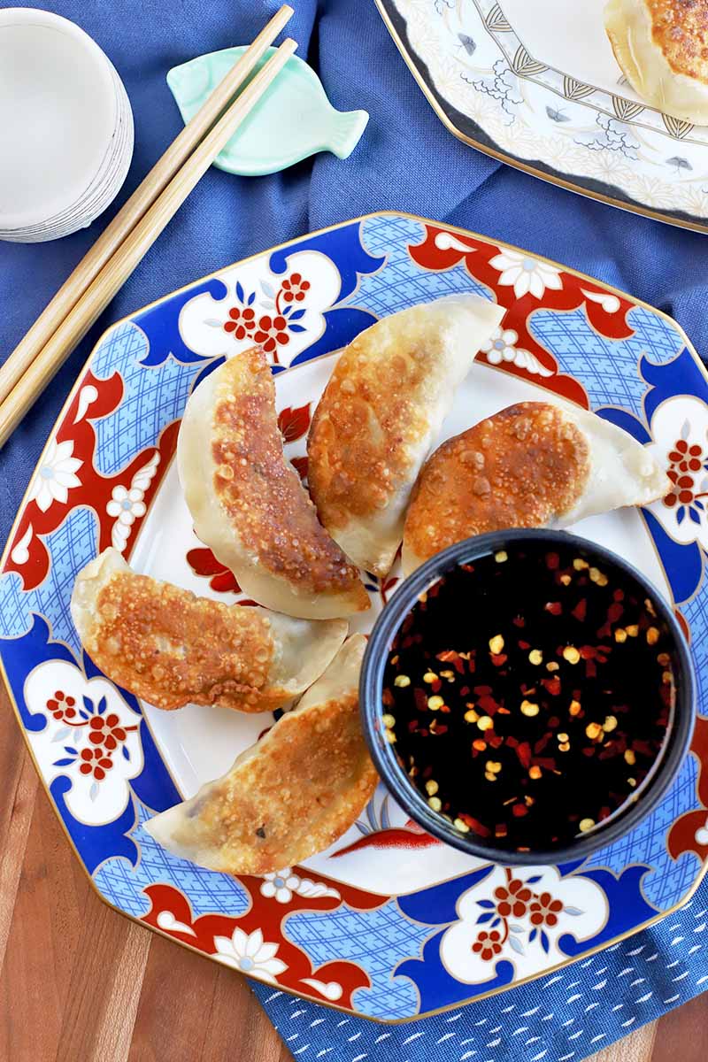 Vertical overhead shot of one white patterned plate and one white, red, and blue patterned plate of homemade pan-fried chicken pot stickers with soy-based dipping sauce, with chopsticks on a blue fish-shaped rest and a small white bowl, on top of a blue cloth on a brown wood table.