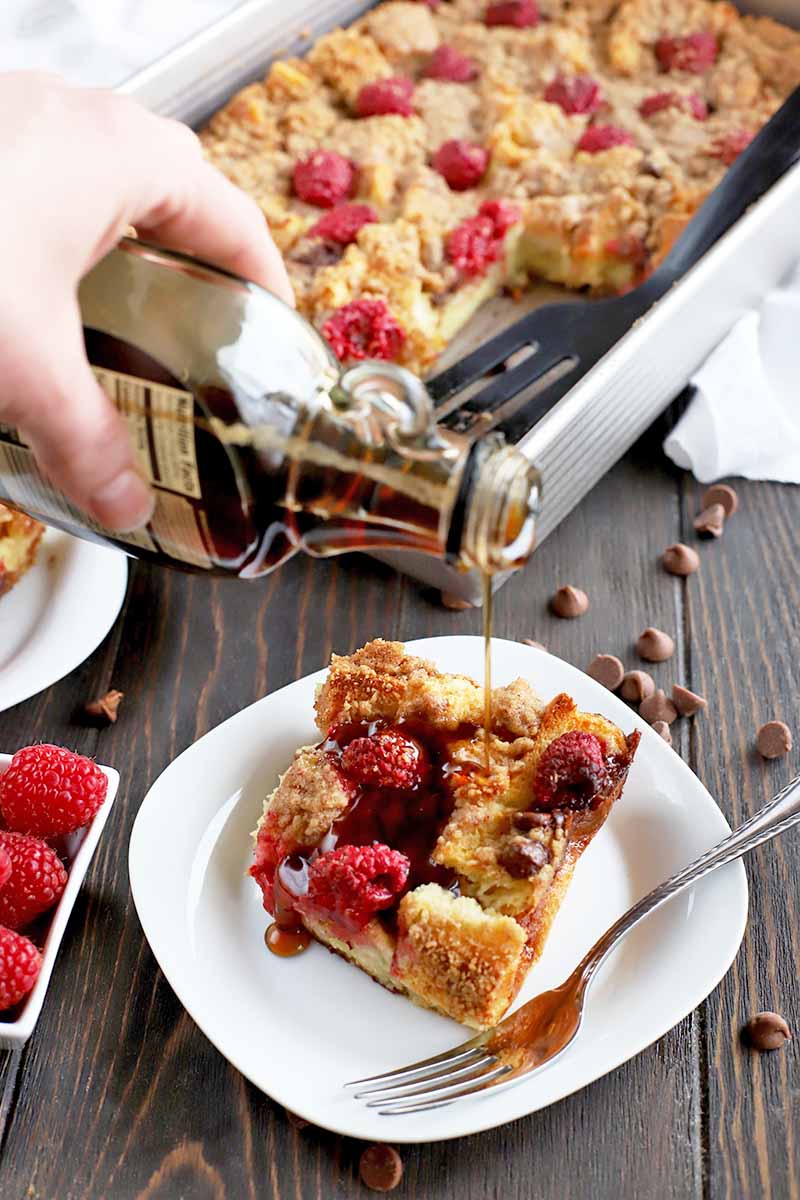 A hand at the left of the frame pours maple syrup from a glass bottle onto a portion of French toast casserole on a plate below, with a fork to the right, and a small dish of fresh raspberries, another plate, a metal baking pan filled with the rest of the casserole, a white cloth, and scattered chocolate chips surrounding the plated portion on a dark brown wood surface.