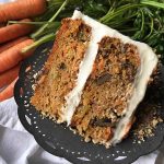 Horizontal image of a slice of carrot cake on a dark plate with fresh vegetables in the background.