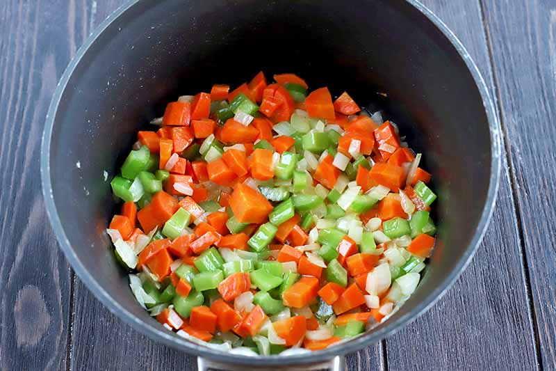 Horizontal overhead shot of chopped orange carrots, green celery, and white onion in the bottom of a nonstick saucepan, on a dark brown wood surface.