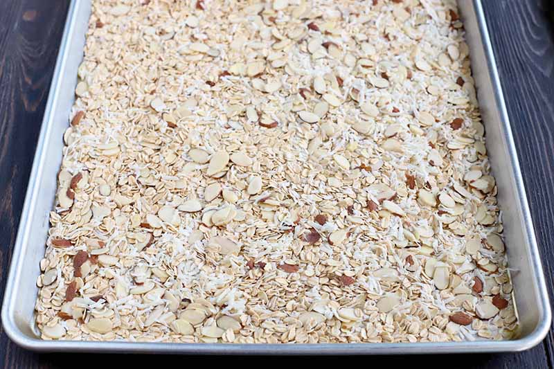 A large rimmed sheet pan of sliced almonds, shredded coconut, and uncooked oats, on a dark brown wood surface.