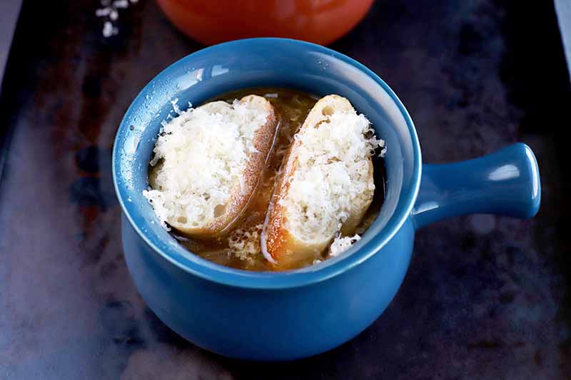 French onion soup in a blue ceramic crock with a handle, topped with baguette slices and melted cheese, on a black metal baking sheet, with a brown ceramic bowl in the background.