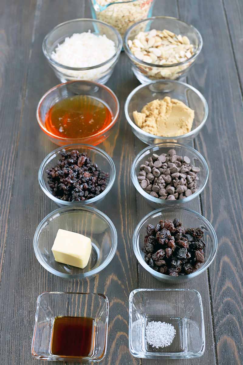 Vertical oblique overhead shot of a small glass measuring pitcher of uncooked oats, eight small round glass bowls of coconut, almonds, honey, brown sugar, dried currants, chocolate chips, butter, and raisins, and two small square glass dishes of vanilla extract and salt.