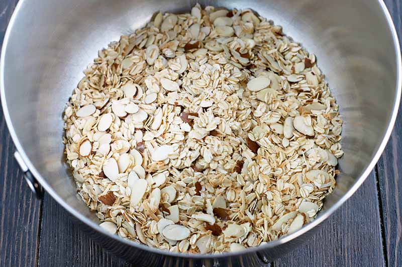 A stainless steel mixing bowl of oats, slivered almonds, and shredded coconut, on a dark brown wood surface.