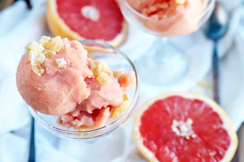 Overhead shot of a glass dessert dish of two scoops of pink sorbet topped with chopped crystallized ginger, with another identical dish to the right, with two spoons and two halves of ruby red grapefruit on a white cloth.
