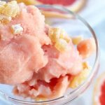 Overhead closely cropped shot of two scoops of pink sorbet in a clear glass dish with crystallized ginger garnish, with two halves of a ruby red grapefruit in soft focus on a white cloth background.
