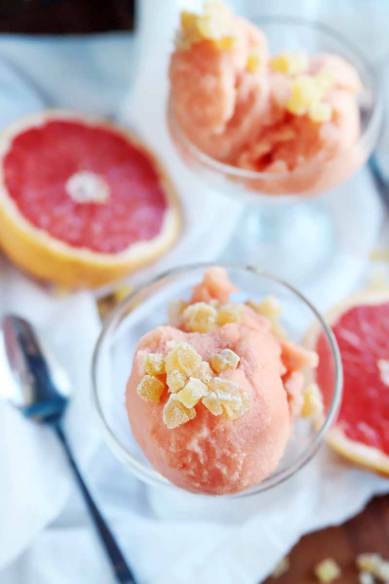 Vertical overhead shot of two glass cups of pink homemade sorbet with candied ginger garnish, on a white cloth with two cut halves of a red grapefruit and a spoon, with scattered ginger on a brown table.