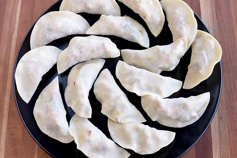 Overhead horizontal shot of sixteen homemade chicken pot stickers in premade wonton wrappers on a black plate ready for frying, on top of a brown wood surface.