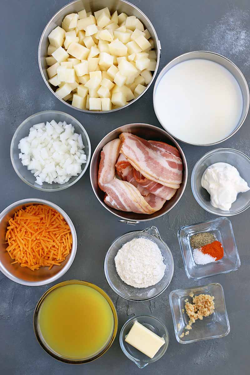 Overhead vertical image of eleven glass and stainless steel dishes of various shapes and sizes containing cubed raw potato, milk, chopped onion, raw bacon, sour cream, spices, minced garlic, chicken broth, shredded cheddar cheese, and flour, on a gray slate surface.