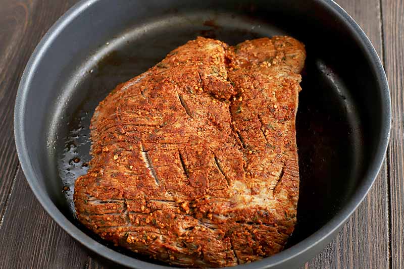 Browned brisket coated in an orange spice rub in a pan, on a dark brown wood surface.
