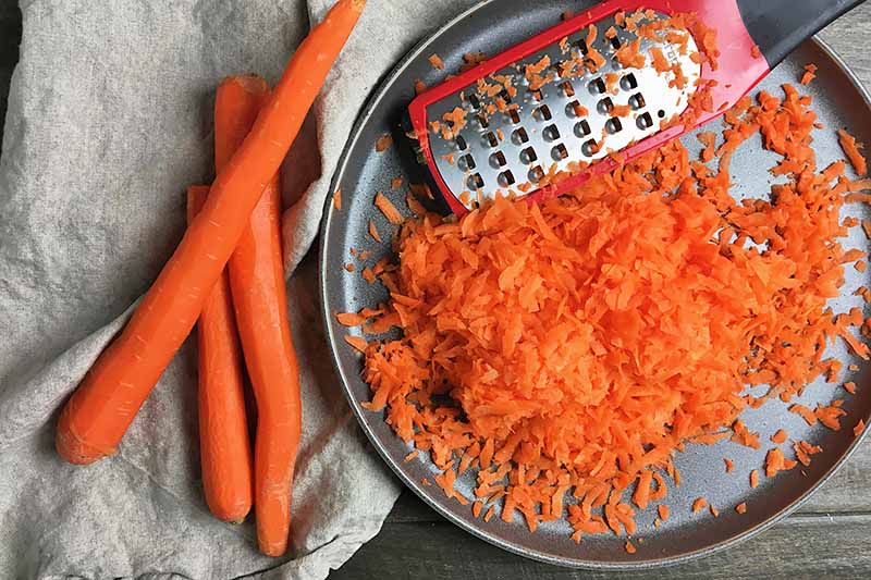 Horizontal image of whole and grated carrots on a plate and towel next to a grater.