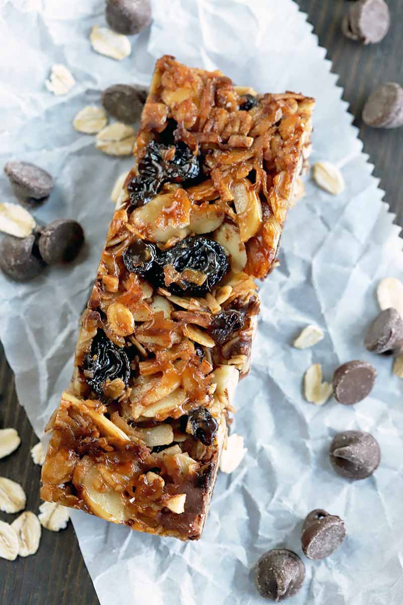 Vertical overhead shot of a homemade granola bar on a white slip of parchment paper, on a dark brown wood surface with scattered chocolate chips and oats.