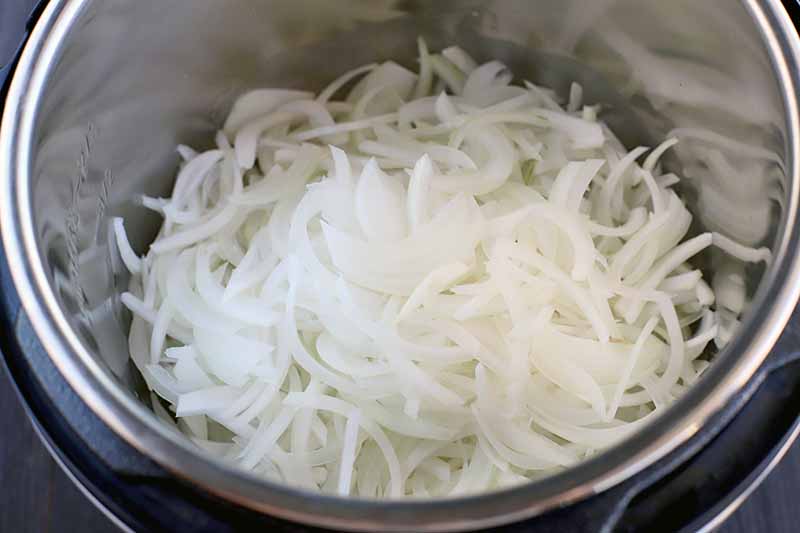 Thinly sliced white onions in a stainless steel pressure cooker insert, on a black background.