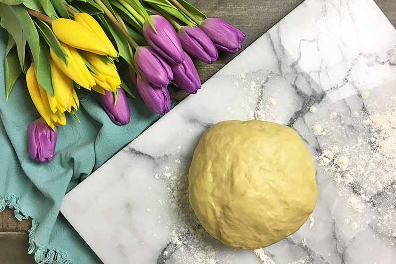 Horizontal image of a bread dough round on a marble board next to a blue towel and flowers.