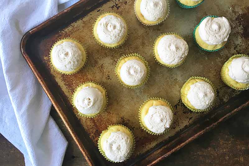 Horizontal image of yellow cupcakes topped with buttercream on a sheet pan next to a white towel.
