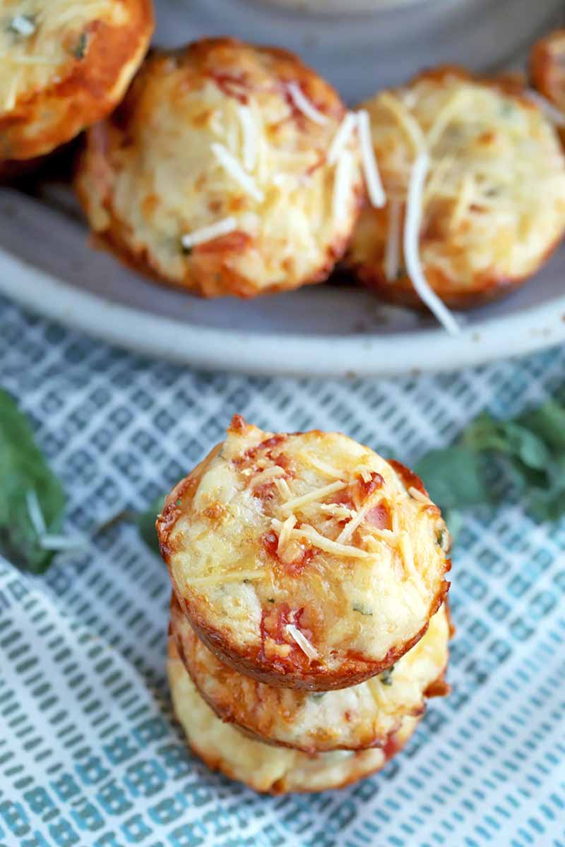 Vertical image of a short stack of three pepperoni and mozzarella cheese savory muffins in the foreground, with more arranged in a circle on a plate in the background, on a blue and white checkered cloth with scattered basil leaves.