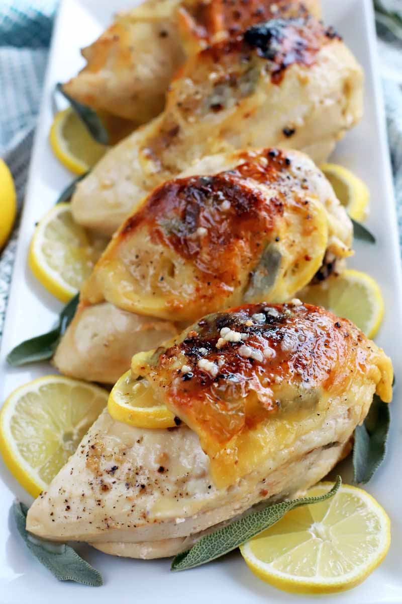 Vertical image of skin-on roast chicken breasts with lemon and sage, on a white ceramic serving platter.