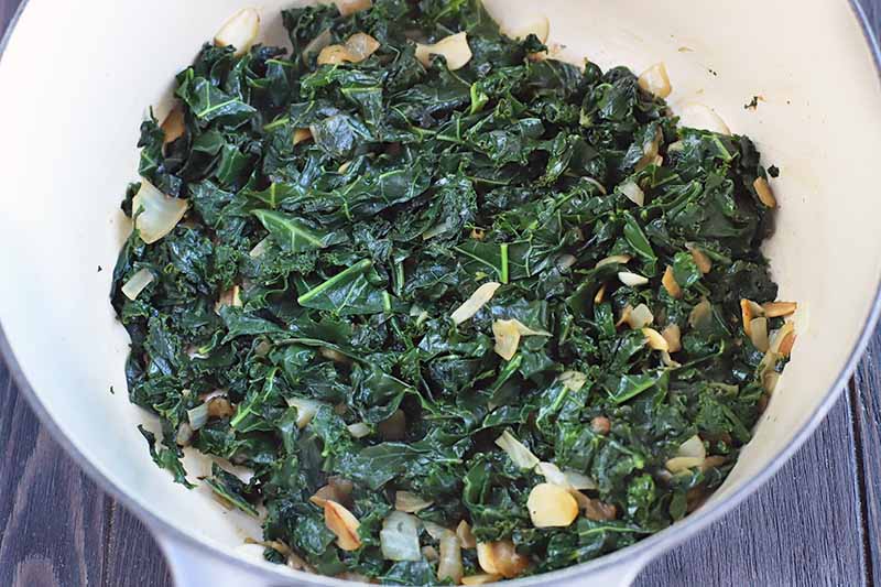 Sauteed kale with garlic and onion in an enameled cast iron cooking pot, on a dark brown wood table.