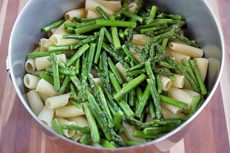 Horizontal shot of rigatoni and thin spears of roasted asparagus in a stainless steel bowl on a wood table.