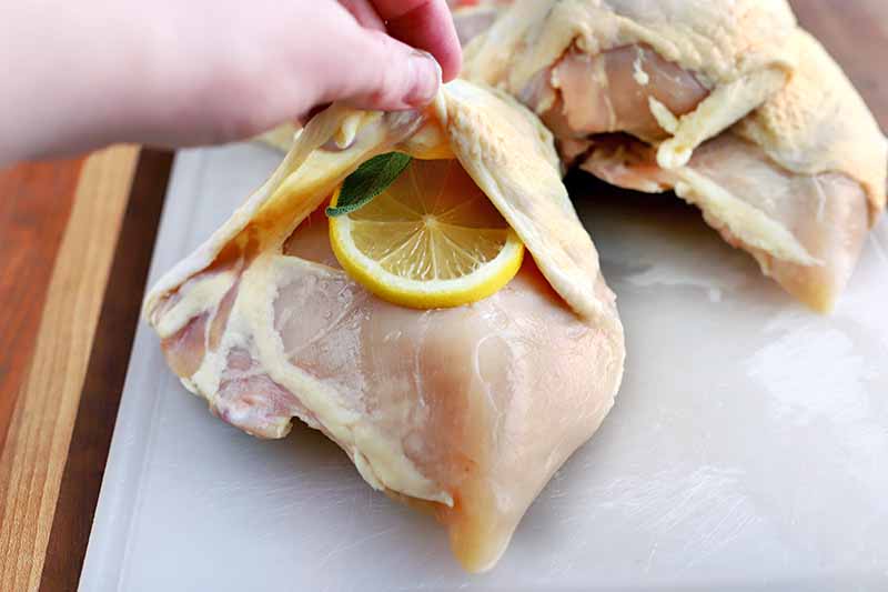 A hand holds up the skin on a piece of chicken and stuffs fresh sage and a slice of lemon inside, on a white cutting board with more poultry towards the top right of the frame, on a wood surface.