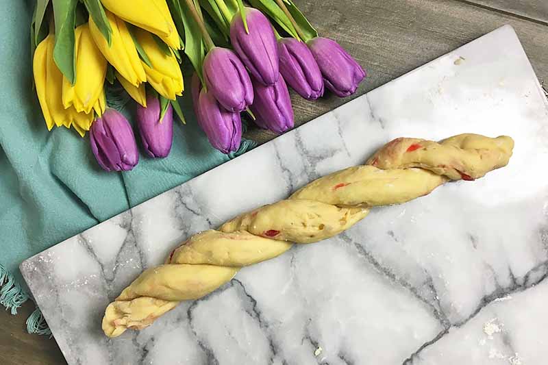 Horizontal image of twisted dough with dried fruit mixed in on a marble board next to yellow and purple flowers.