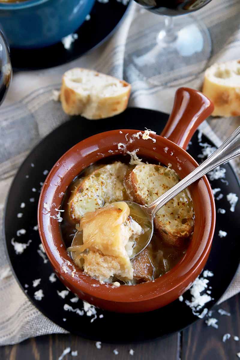 Vertical overhead shot of a brown crock of French onion soup with a spoon, on a black plate with scattered grated cheese, on a striped gray cloth with a glass of wine and a blue crock on a plate in the background, with two slices of baguette in between, on a dark brown wood table.