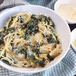 A white bowl of noodles and greens on a blue and white cloth, with a fork, two small white bowls of citrus zest and grated Parmesan cheese, and a whole lemon, on a dark brown wood surface.