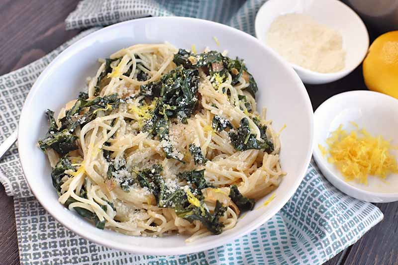 A white bowl of noodles and greens on a blue and white cloth, with a fork, two small white bowls of citrus zest and grated Parmesan cheese, and a whole lemon, on a dark brown wood surface.