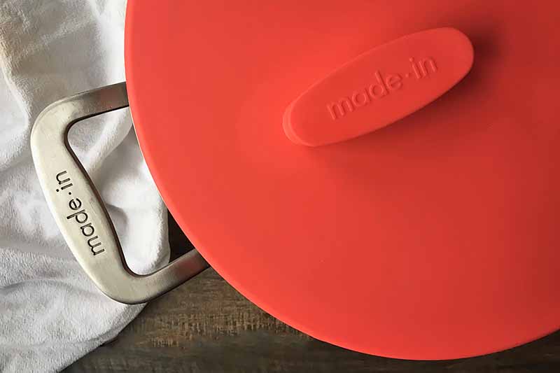 Horizontal image of a lid on top of a pan with a handle next to a towel.
