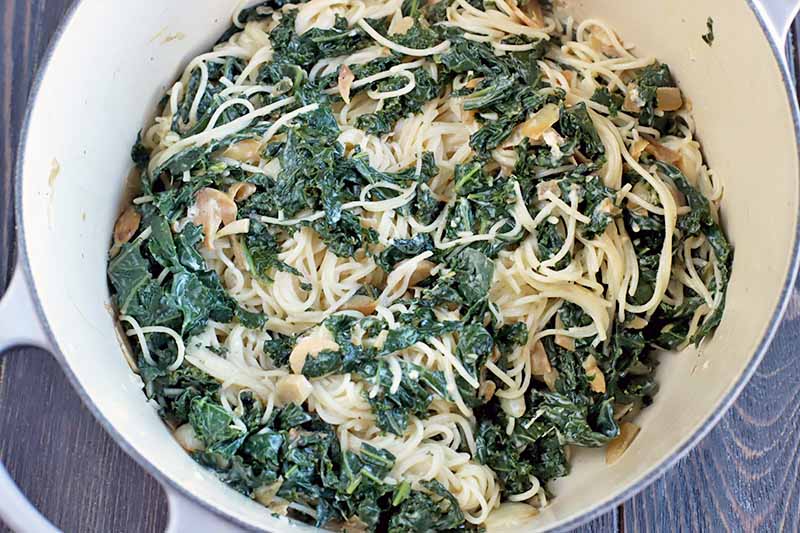 Cooked leafy greens, noodles, onion, and garlic, in the bottom of a cream-colored enameled cast iron pot that is light blue on the outside, on a dark brown wood surface.