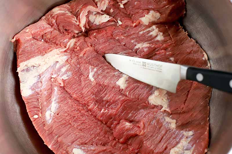 A paring knife is used to slice slits into a raw piece of beef, in a large stainless steel bowl.