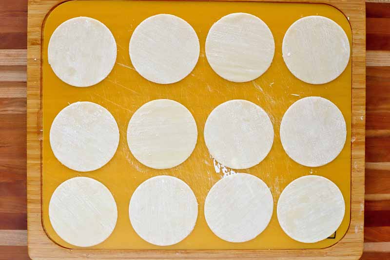 Overhead shot of twelve round wonton wrappers arranged in three rows on a yellow plastic and wood cutting board that is lightly dusted with flour, on top of a striped brown and wood beige table.
