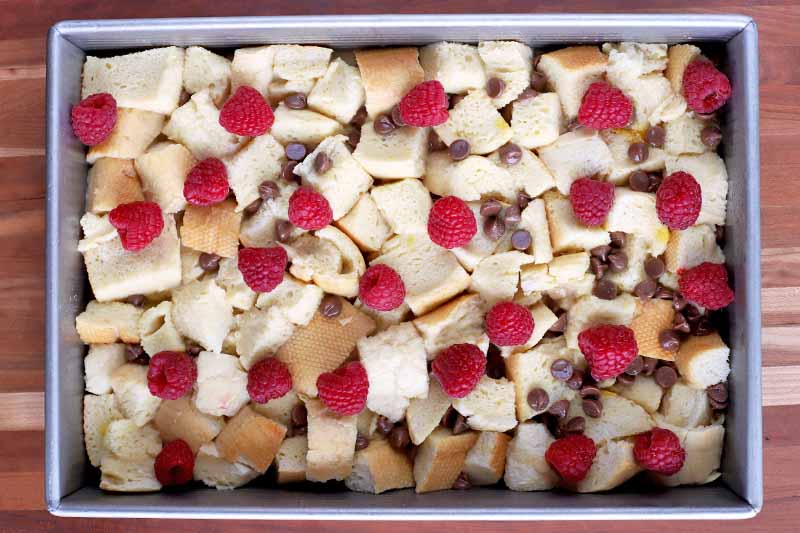Overhead shot of a metal baking pan of bread cubes soaked in egg, chocolate chips, and fresh raspberries, on a striped wood surface.
