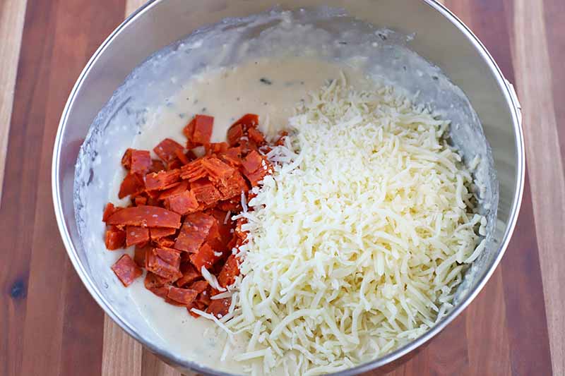 Overhead horizontal shot of a stainless steel bowl of savory muffin batter with a pile of chopped pepperoni slices and a larger pile of shredded mozzarella cheese on top, on a beige and brown striped wood surface.
