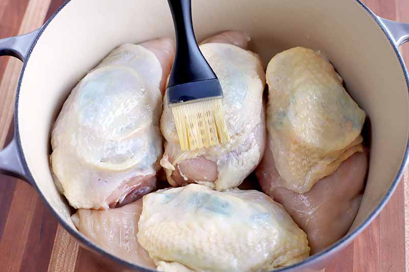 A pastry brush is used to apply a thin layer of oil to the skin on four pieces of bone-in chicken breast, in a light blue and cream-colored enameled cast iron Dutch oven, on a wood surface.
