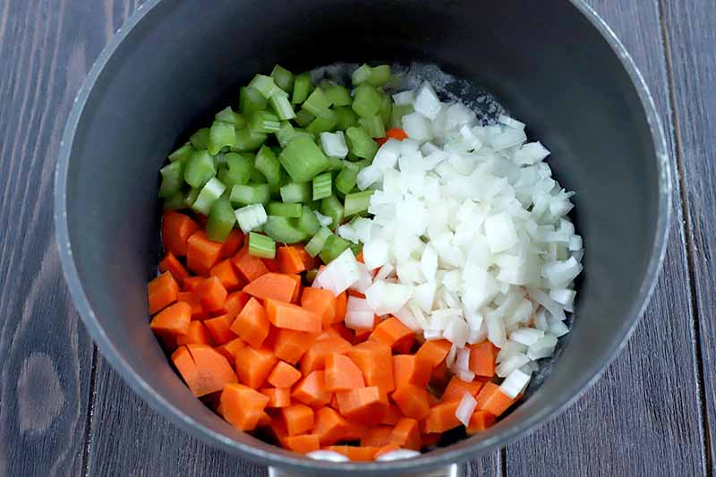 Overhead shot of a nonstick saucepan with three piles of raw chopped vegetables at the bottom, with onions and celery taking up one-quarter each, and carrots taking up half of the bottom of the pan, on a dark brown wood table.
