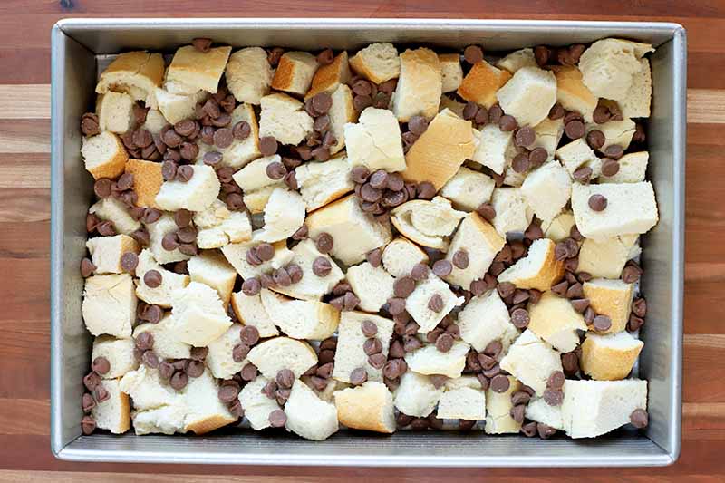 Overhead shot of bread cubes topped with chocolate chips in a rectangular metal baking pan, on a wood surface.