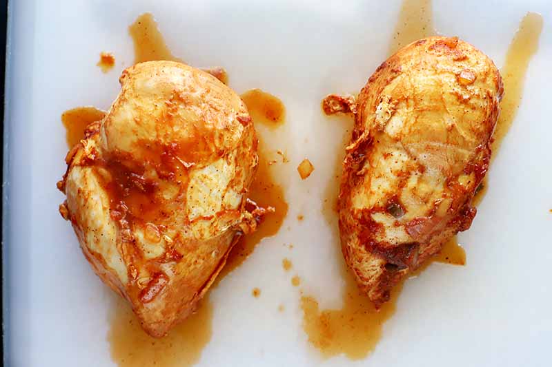 Horizontal image of two cooked chicken breasts covered in a red sauce.