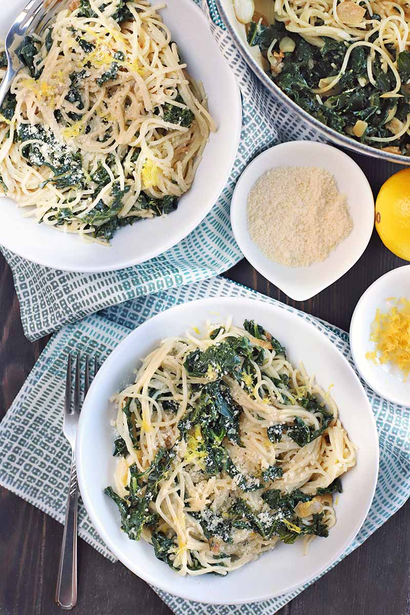 Overhead vertical shot of two shallow bowls of cooked noodles and greens, on two blue and white cloth napkins with a fork, with two small white bowls of citrus zest and grated parmesan, a whole lemon, and a stainless steel bowl of more of the dish, on a dark brown wood table.