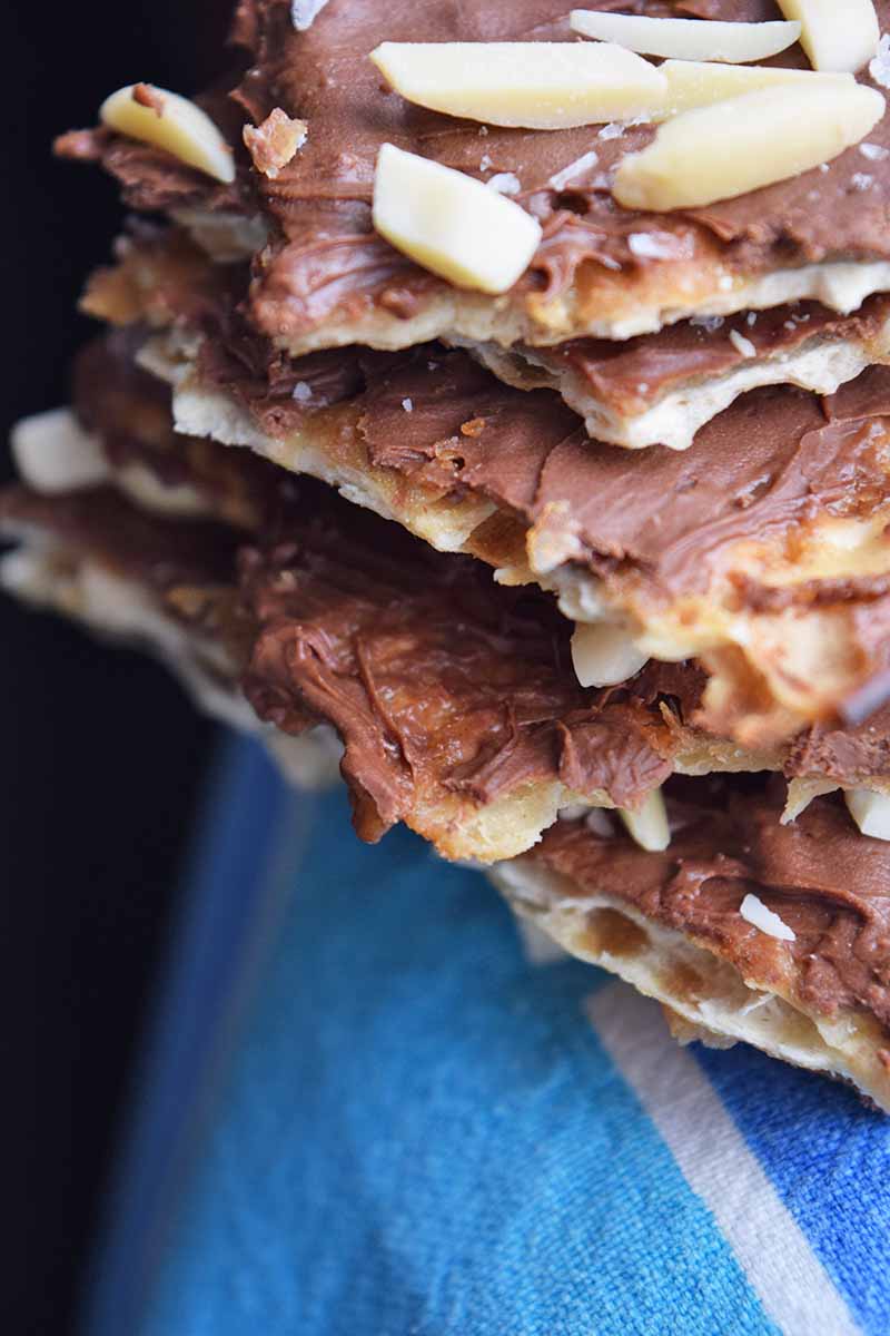 Vertical closeup image of a stack of chocolate-covered almond toffee matzoh on a white and blue striped cloth, with a black background.