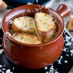 Closeup of a brown glazed ceramic crock of French onion soup topped with toasted baguette slices and melted cheese, on a black plate with scattered bits of grated Swiss, on a gray cloth with bread and a wine glass.