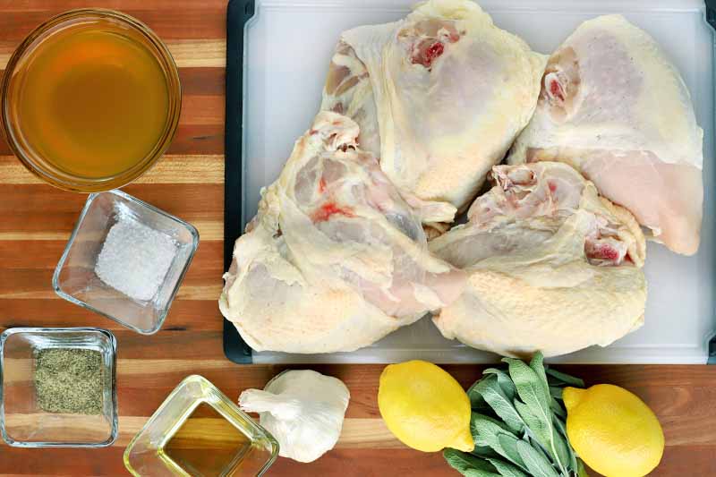 Overhead shot of three raw chicken breasts with the bone and skin on a black and white cutting board, on a wood surface with a small glass bowl of stock, small square glass bowls of salt, pepper, and olive oil, a head of garlic, two lemons, and a small bunch of fresh sage.