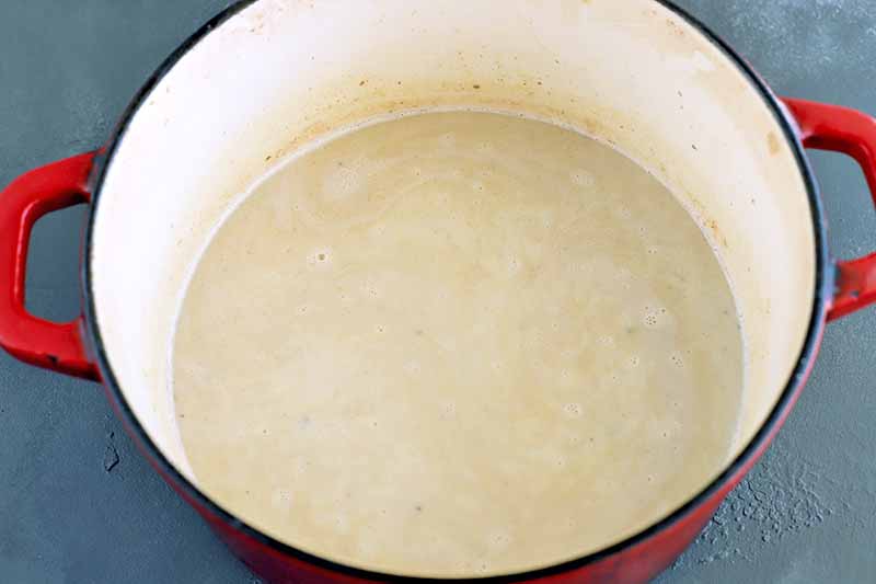 Potato soup filled a red and cream-colored enameled Dutch oven with two handles, on a gray slate surface.